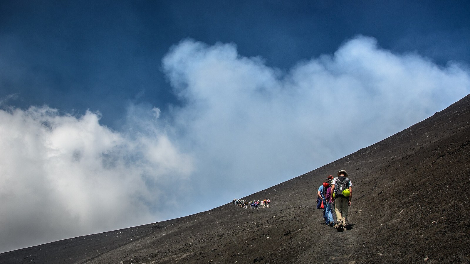 Hiker ascending Mount Etna on a volcanic gravel slope with cloud below